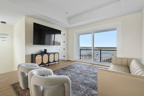 Una sala de estar moderna con un televisor de pantalla plana, un sofá, dos sillones, una consola y un amplio ventanal corredero que da a un balcón con vistas al agua.