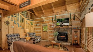 LâintĂ©rieur chaleureux dâun chalet avec une cheminĂ©e, des murs en bois, un escalier et une pancarte sur laquelle on peut lire : « Câest ici que la famille et les amis se rĂ©unissent ».