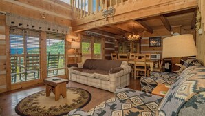 LâintĂ©rieur dâun chalet confortable, avec un canapĂ© Ă carreaux, un pouf Ă motifs, une table basse en bois et un coin repas comprenant une table et des chaises.