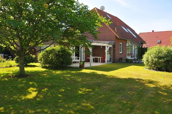 Enjoy outdoor dining or relaxation on the covered patio surrounded by lush gardens and a large tree.