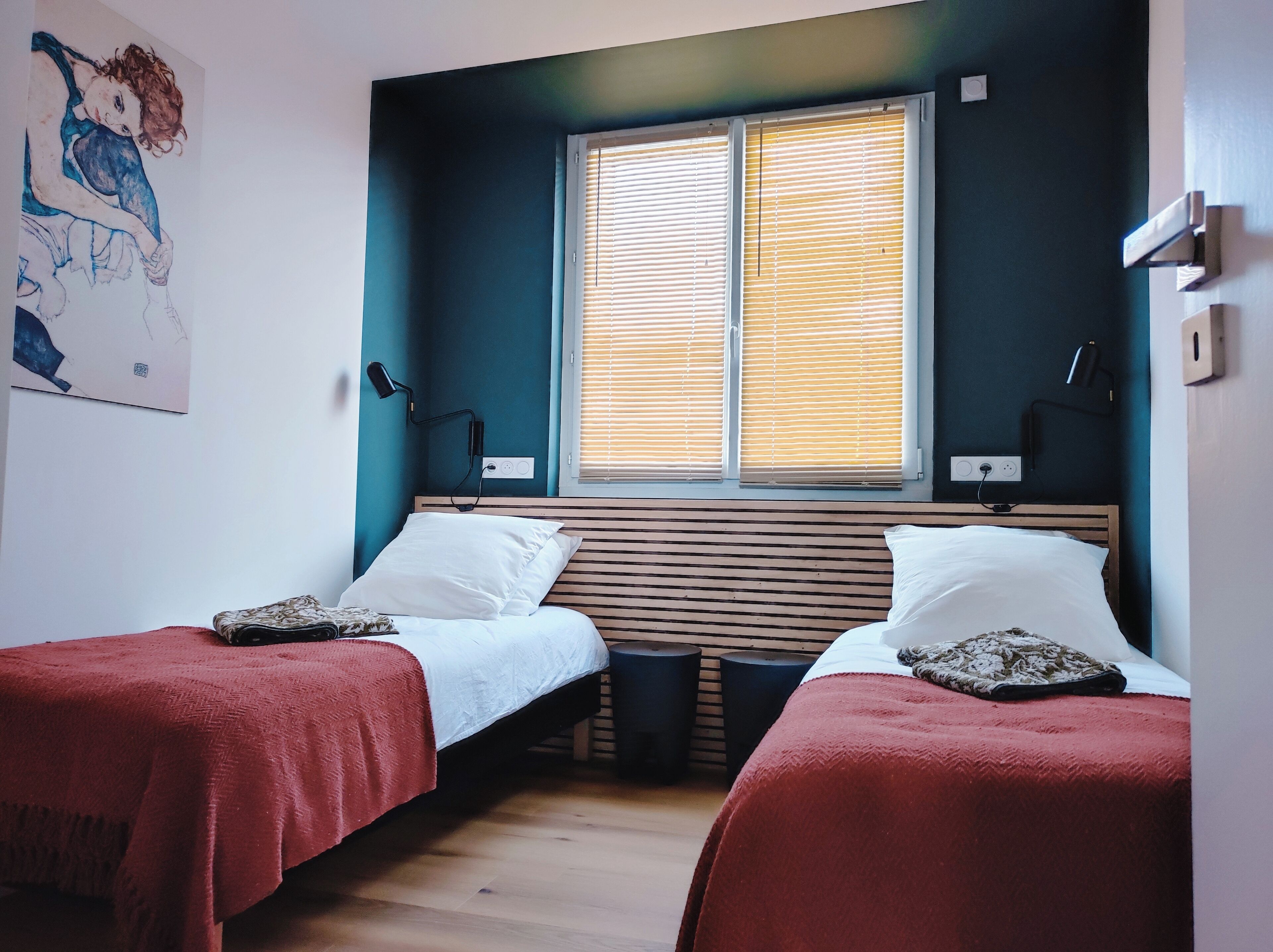 A room with two beds, a window with blinds, a wall-mounted lamp, and a painting.