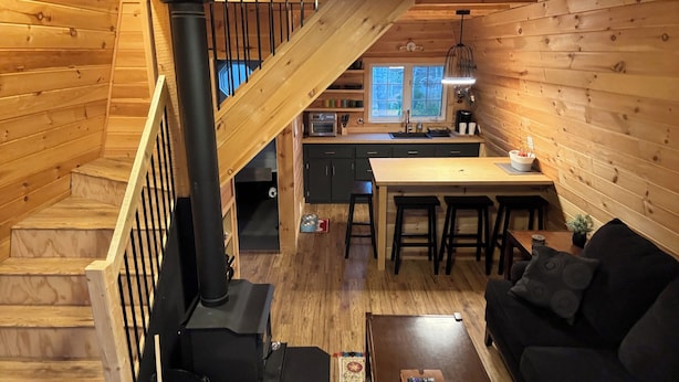 A cozy cabin interior with a wooden staircase, a central fireplace, a kitchen area, and a living room furnished with a sofa and a coffee table. - Unplug and relax in this quiet retreat on the shores of Tracadie Bay (Donaldston)