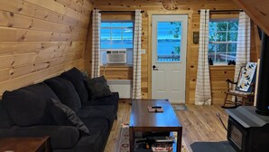 A cozy cabin interior with a wooden interior, a black sofa, a small table, a fireplace, and a view through the door. - Unplug and relax in this quiet retreat on the shores of Tracadie Bay (Donaldston)