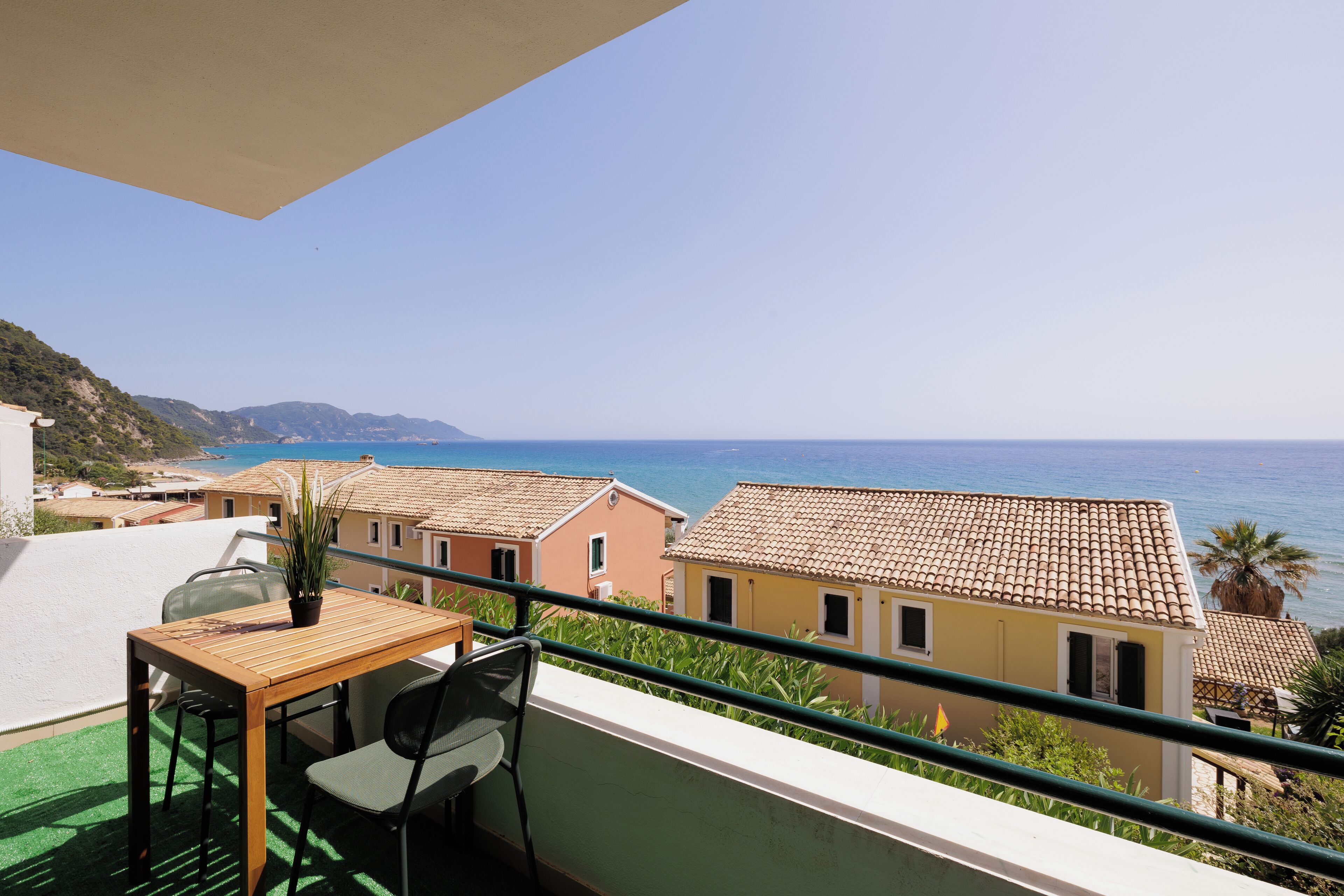 A balcony with a wooden table and chairs, overlooking a coastal town with terracotta-roofed buildings and the sea.