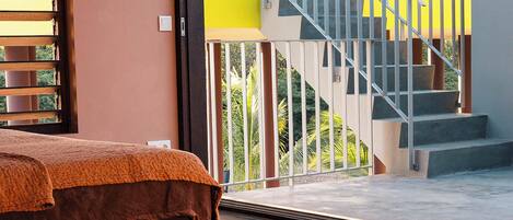 A bedroom with a bed, a window with blinds, and a view of a staircase leading to an outdoor area.