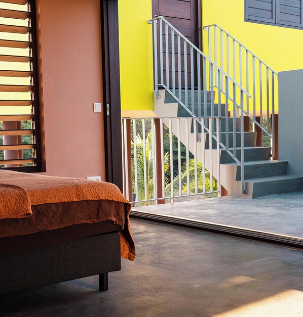 A bedroom with a bed, a window with blinds, and a view of a staircase leading to an outdoor area.