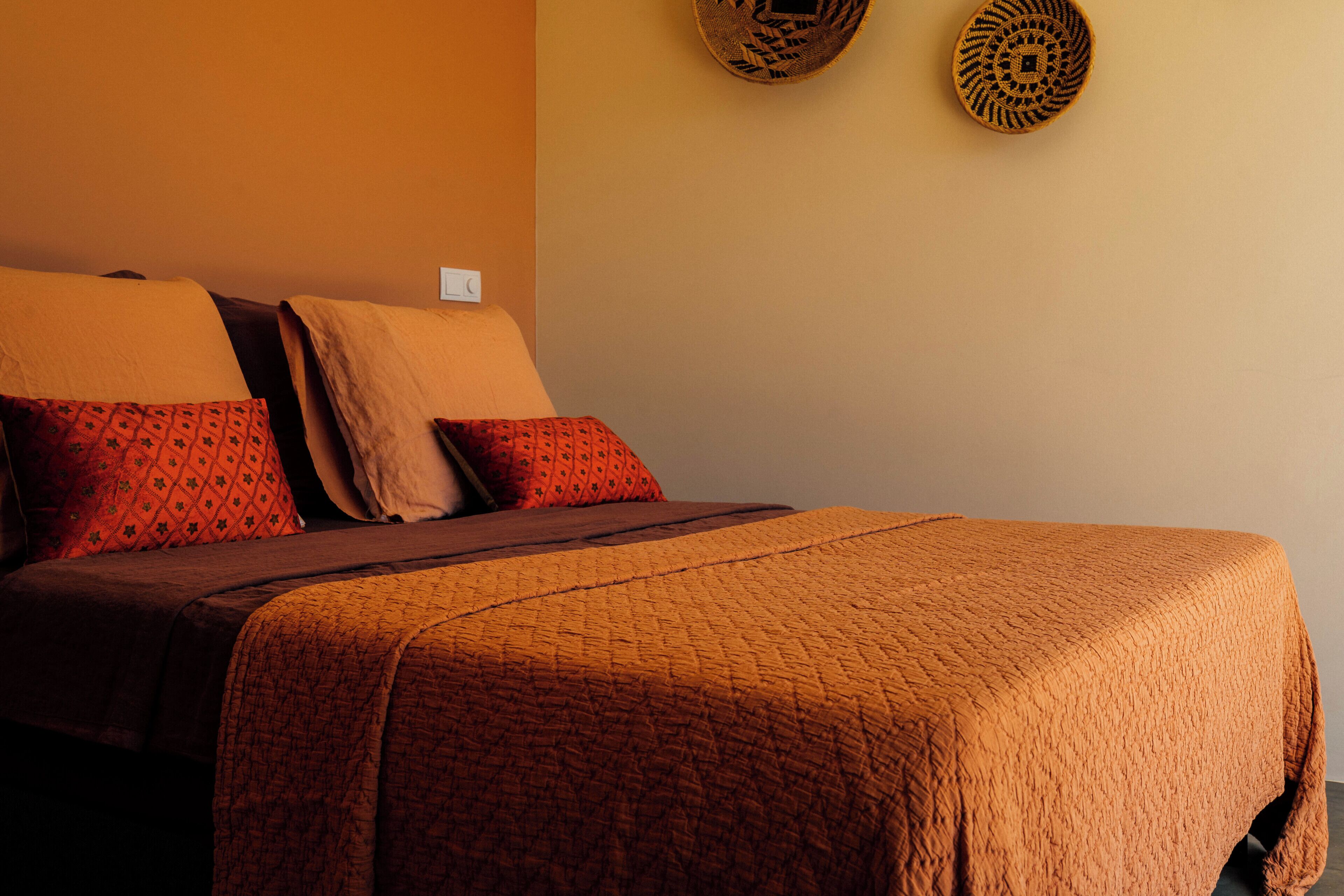 A bed with a brown quilt and red pillows, a bedside table, and a wall-mounted light switch.