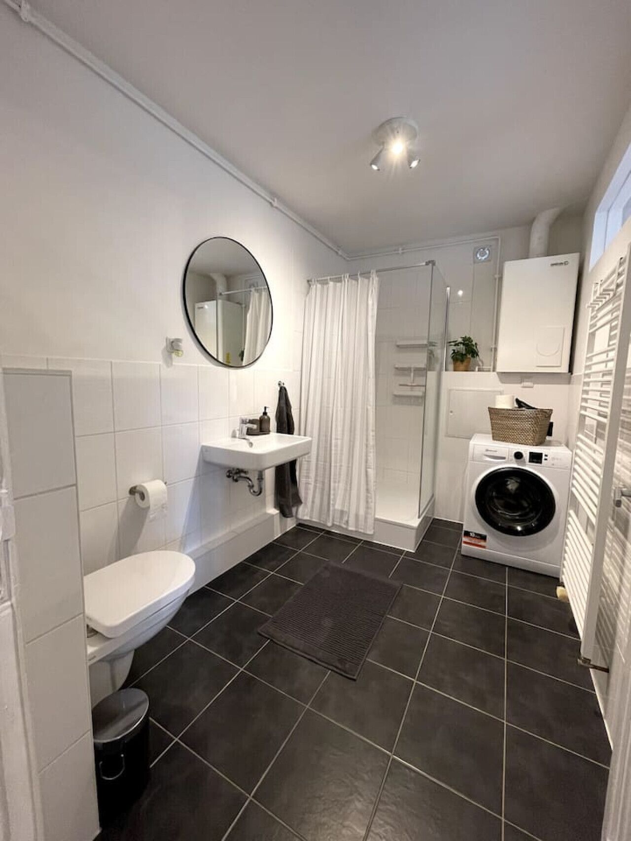 Ein modernes Badezimmer mit Toilette, Waschbecken, Spiegel, Dusche und einer integrierten Waschmaschine.
