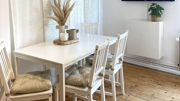 Un coin repas avec une table blanche, quatre chaises, un vase contenant des plantes séchées, une bougie et un téléviseur fixé à un mur blanc.