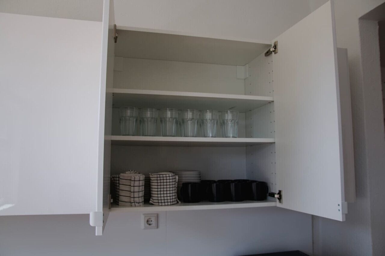 Un mueble blanco con varios estantes, que contiene vasos apilados, servilletas a cuadros y recipientes negros.