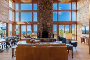 A spacious living room with a stone fireplace, large windows, and a view of the mountains. - Skytop Chateau at The Summit of Beech (Banner Elk)