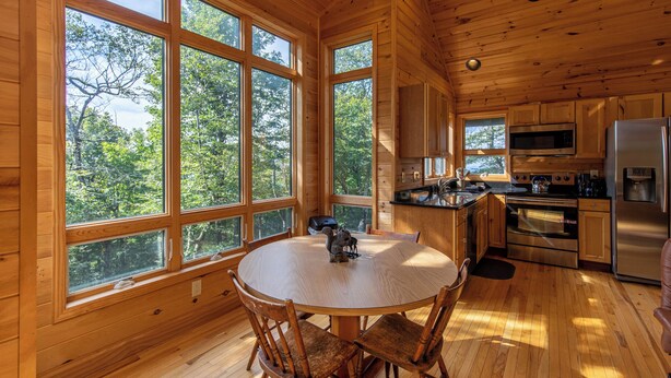 A wooden cabin with a round dining table and chairs, large windows, a kitchen with modern appliances, and a view of the forest. - Chalet at The Summit of Beech (Banner Elk)