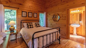 A wooden cabin bedroom with a bed, a bedside table with a lamp, a window with curtains, and a bathroom with a sink. - Chalet at The Summit of Beech (Banner Elk)