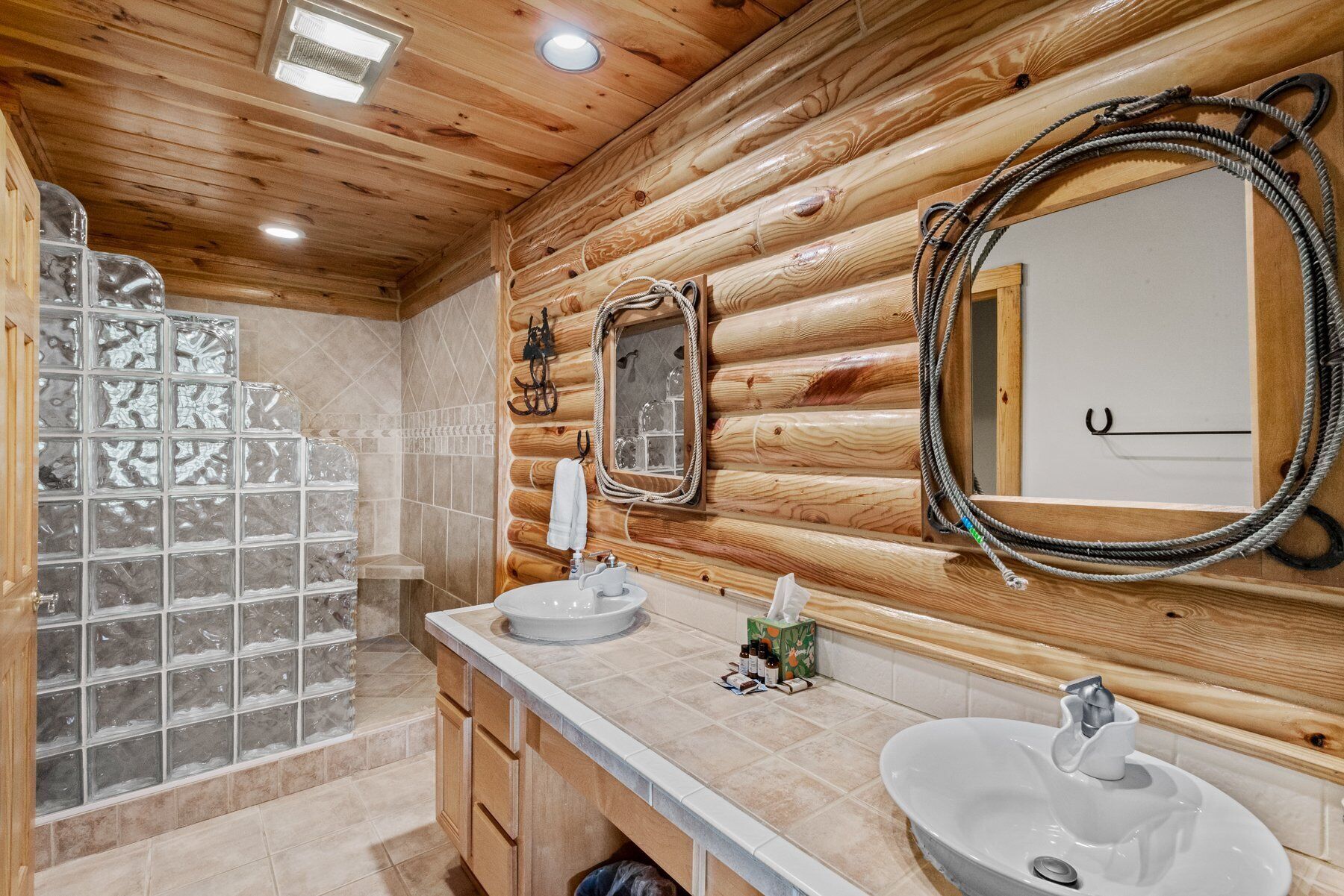 A bathroom with wooden log walls, two sinks, a mirror, and a glass shower enclosure.