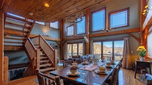 Un vaste espace dédié à la salle à manger, avec une longue table en bois dressée pour le repas, un escalier menant aux niveaux supérieurs et de grandes fenêtres offrant une vue sur l’extérieur.