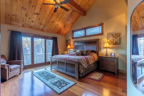 A bedroom with a wooden ceiling, a large bed, a nightstand, a lamp, and a window with a view of trees. - Elk Island at River Run (Banner Elk)