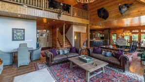A spacious living area with a high ceiling, wooden beams, and a mezzanine level. There are two sofas with plaid cushions, a coffee table, and a chandelier. - A High Country Retreat (Boone)