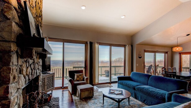 A spacious living room with a stone fireplace, a blue sofa, and a large sliding glass door leading to a balcony with a view of the mountains. - Hill Station (Banner Elk)