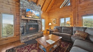 L’intérieur chaleureux d’un chalet avec une cheminée en pierre, des murs en bois, un canapé en cuir et une grande fenêtre.