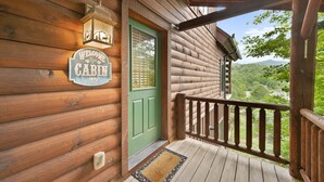 Une cabane en bois avec une porte verte, une pancarte indiquant « Bienvenue » et un balcon doté d’une balustrade en bois.