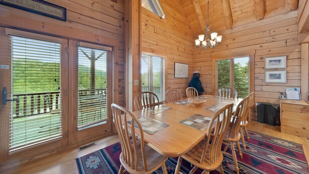 L’intérieur d’un chalet en bois comprend une table à manger, des chaises, un lustre et de grandes fenêtres équipées de stores.