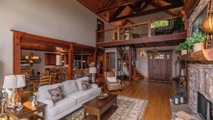 A spacious living area with a high ceiling, wooden beams, a fireplace, and a dining area with a kitchen in the background. - Haven at Headwaters (Elk Park)