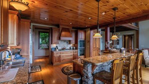 A spacious kitchen with wooden cabinets, granite countertops, and a large island with bar stools. - Haven at Headwaters (Elk Park)