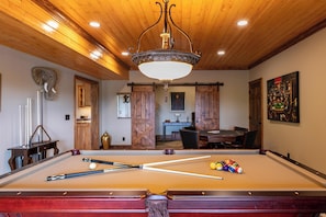 Une table de billard avec des billes, une queue de billard et une autre table de billard dotée d’un plafond en bois et d’un lustre.