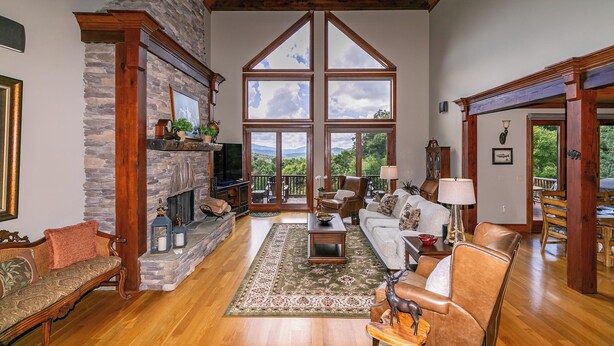 A spacious living room with a stone fireplace, wooden beams, and large windows offering a view of the outdoors. - Haven at Headwaters (Elk Park)