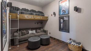 Een kamer met een stapelbed met de aanduiding 'Kids Loft', een bank, twee voetenbankjes, een gepresenteerde poster van een haai, en een mand met spullen.