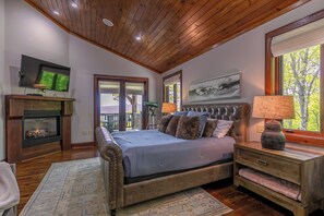 A bedroom with a fireplace, a television, a bed with a headboard, a nightstand, and a window with a view. - Sanctuary at Eagles Nest (Banner Elk)