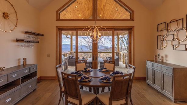 Un espace repas avec une grande table en bois, des chaises et un lustre. De grandes fenêtres offrent une vue sur l’extérieur, et la pièce est agrémentée de poutres en bois au plafond.