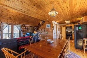 Interior de una cabaña de madera con una mesa de comedor, sillas, un sofá, una cocina con refrigerador y una escalera que conduce a un altillo.