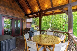 Een terras buiten met een houten tafel en stoelen, een barbecue en een overdekt gedeelte met uitzicht op bomen.