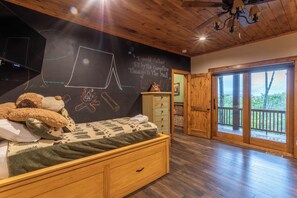 Een gezellige slaapkamer met een houten bed, een prikbordwand met tekeningen, een dressoir en een groot raam met uitzicht op bomen.