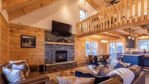 A cozy log cabin interior with a stone fireplace, leather sofas, a dining area, and a kitchen with a large window. - Ursa Minor (Banner Elk)