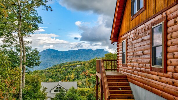 A log cabin with a wooden staircase leading to a deck, surrounded by trees and mountains. - Ursa Minor (Banner Elk)