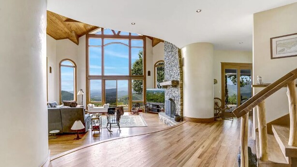 A spacious living area with large windows, a stone fireplace, and a wooden staircase. - Sunrise to Sunset (Hayesville)