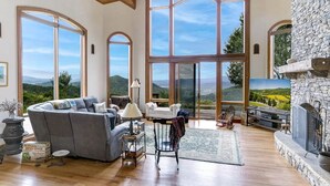 A spacious living room with a large fireplace, a stone wall, and a scenic view through large windows. - Sunrise to Sunset (Hayesville)