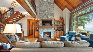 Amplia sala de estar con chimenea de piedra, vigas de madera, una escalera y ventanas grandes que ofrecen vistas al exterior.