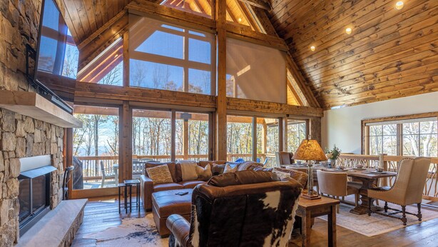 A spacious living area with a stone fireplace, leather furniture, and large windows offering views of the surrounding trees. - Casa Tres Cuervos at Eagles Nest (Banner Elk)