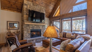 A cozy living room with a stone fireplace, leather furniture, and a large window offering a view of trees. - Casa Tres Cuervos at Eagles Nest (Banner Elk)