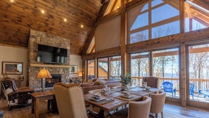 A spacious living room with a stone fireplace, wooden ceiling, and large windows offering a view of the outdoors. - Casa Tres Cuervos at Eagles Nest (Banner Elk)