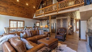 A spacious living room with a high ceiling and wooden beams, a cozy fireplace, and a dining area with a kitchen in the background. - Casa Tres Cuervos at Eagles Nest (Banner Elk)