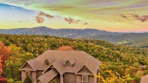 A house with a steeply pitched roof and a small porch, surrounded by dense forest and hills in the background. - Sugar Sky (Banner Elk)