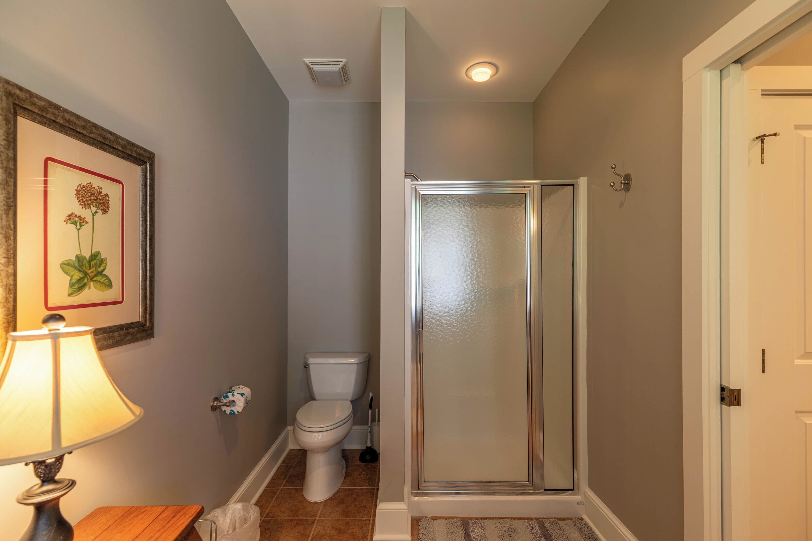 A bathroom with a toilet, a shower with a glass door, a framed picture on the wall, and a floor lamp.