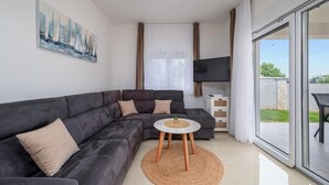 Uma sala de estar moderna com um sofá modular cinza escuro, uma mesa redonda pequena com uma planta, uma televisão de tela plana e uma porta de vidro deslizante que dá para uma área externa.