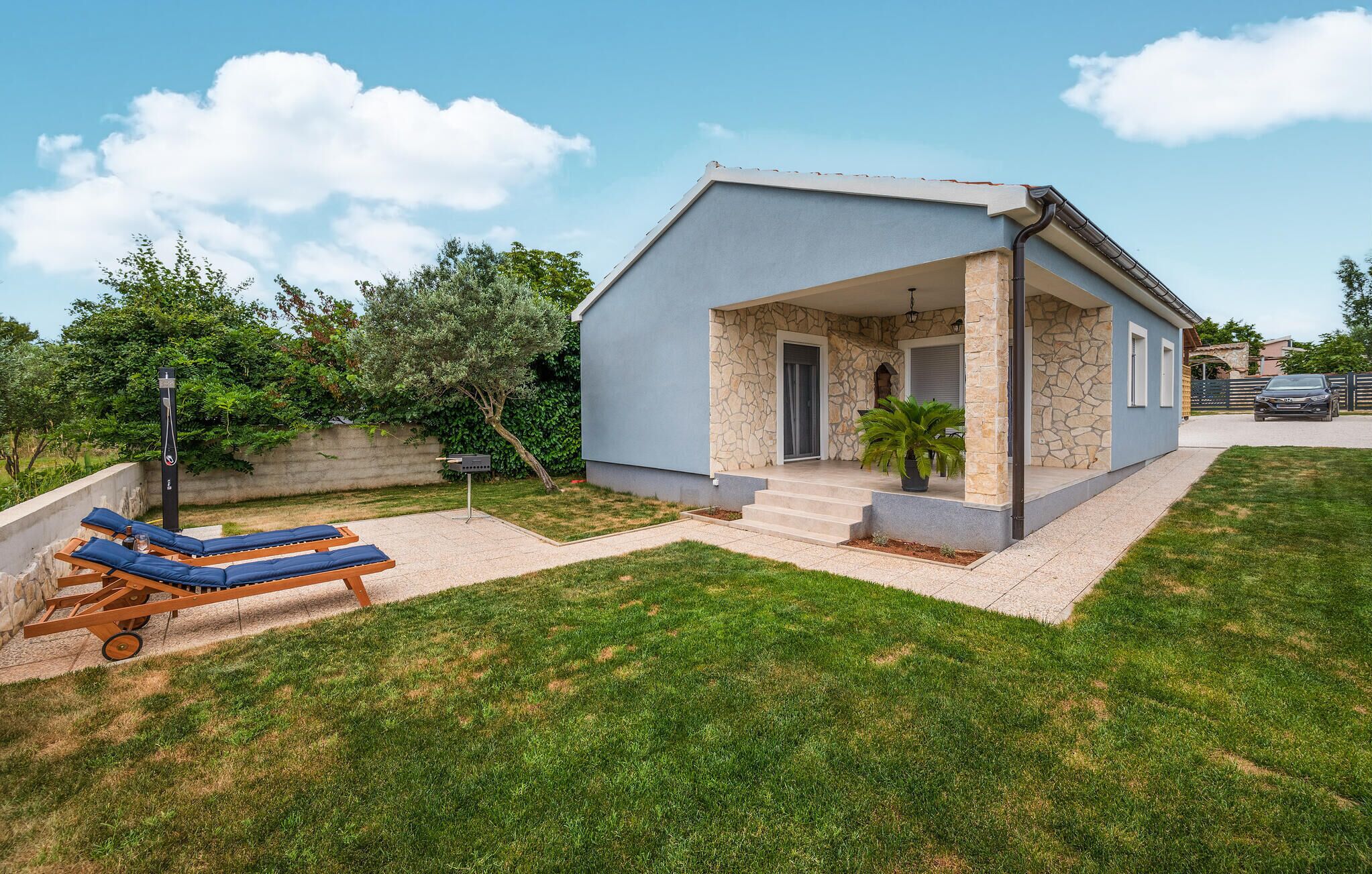 Une maison moderne avec une façade en pierre, un porche abrité et une pelouse agrémentée de chaises longues.