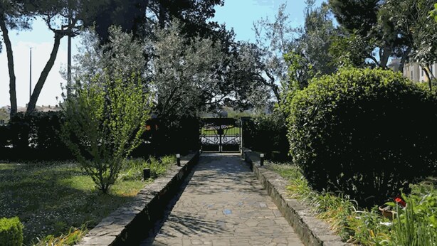 Un chemin de pierre mène à une entrée fermée par un portail, qui est elle-même entourée d’une végétation luxuriante et d’arbres.