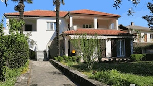 Une maison à deux étages, avec un toit recouvert de tuiles rouges, un chemin de pierre menant à l’entrée et un petit jardin agrémenté de diverses plantes.